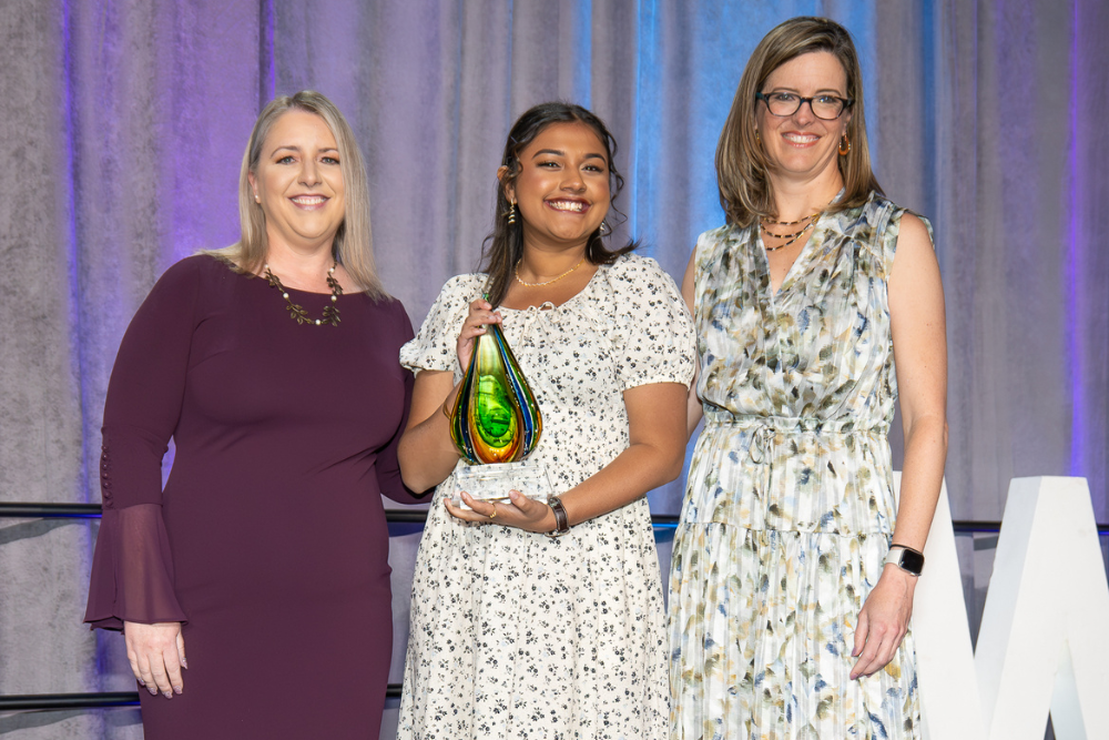 Three women stand together on a stage with purple and blue lighting in the background. The woman in the center, wearing a white dress with small floral print, holds a green teardrop-shaped glass award and smiles broadly. She is flanked by two other women - one in a burgundy dress on the left and another in a light-colored floral dress with glasses on the right.