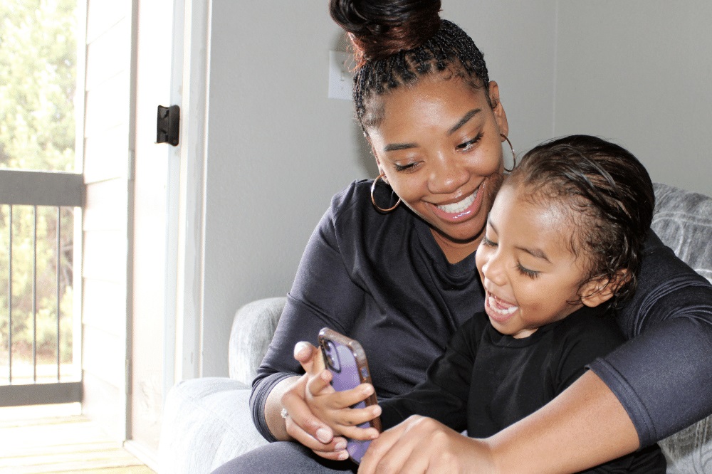 A young Black woman with a child on her lap smiles while looking at her phone