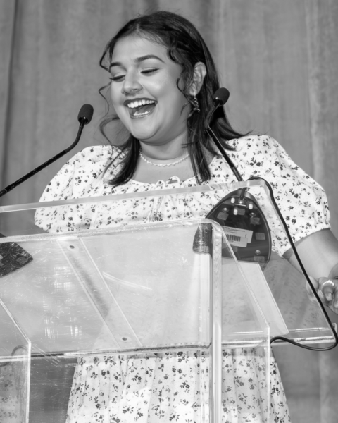 Gitanjali Rao in a white floral print dress stands at a clear acrylic podium, speaking into a microphone with a joyful expression. She appears to be giving an acceptance speech, with curtains visible in the background. The black and white photograph captures her mid-speech, smiling warmly as she addresses the audience.