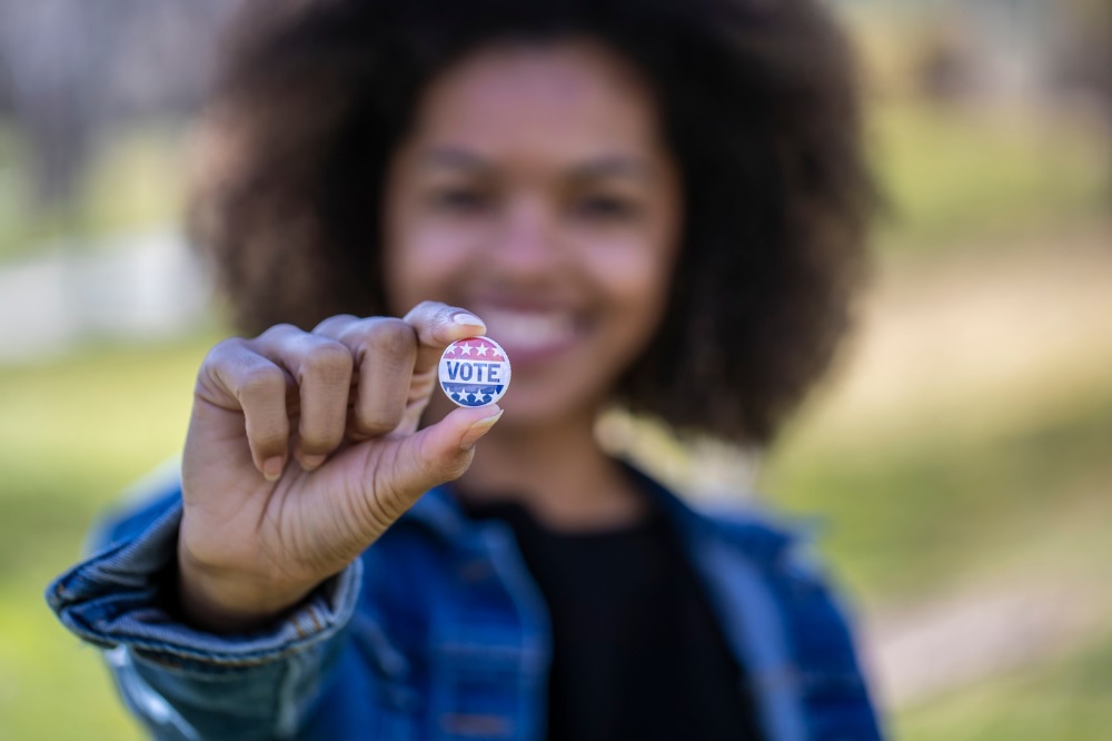 A young smiling woman holds up a button that says I voted.