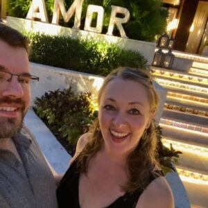 A smiling couple taking a selfie at night in front of illuminated stairs and a sign that reads "AMOR." The woman on the right has blonde hair and is wearing a black dress, while the man on the left has a beard, glasses, and is wearing a button-up shirt.