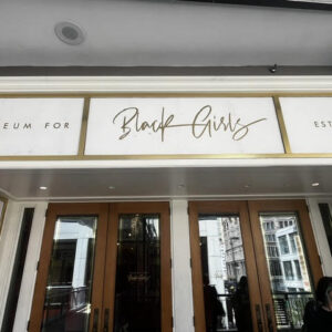 The entrance to 'Museum for Black Girls' established in 2019. The name is displayed in three white panels with gold frames above glass double doors: 'MUSEUM FOR' on the left, 'Black Girls' in script lettering in the center, and 'EST. 2019' on the right.