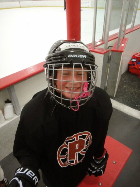 A young girl in full ice hockey gear smiles from the bench area inside an ice rink. She wears a black jersey with a red and white "P" logo, black gloves, and a helmet labeled "DELANEY LINDNER." The ice rink and hockey equipment are visible in the background.