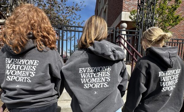 Three women stand outdoors with their backs to the camera, wearing matching dark gray hoodies that read "EVERYONE WATCHES WOMEN'S SPORTS." in bold white lettering. They are standing on steps in front of a gated building under a bright blue sky.