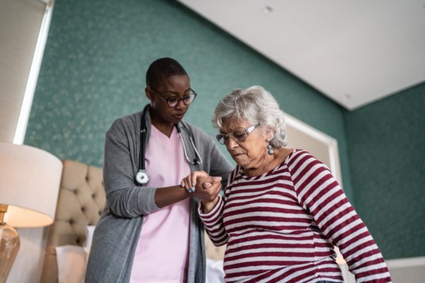 A Black woman who is a home care worker helps an older white woman by holding her hand