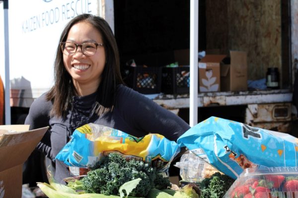 An Asian woman stands in front of a truck and a table full of fresh vegetables