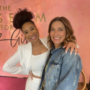 Two women pose together smiling at the Museum for Black Girls. On the left, Mercedes Howard ('Ya Girl Cedes') wears a white two-piece outfit with her hair in a high bun. On the right, Renee Ferrufino wears a denim jacket. They stand in front of a pink wall with part of the museum's logo visible behind them