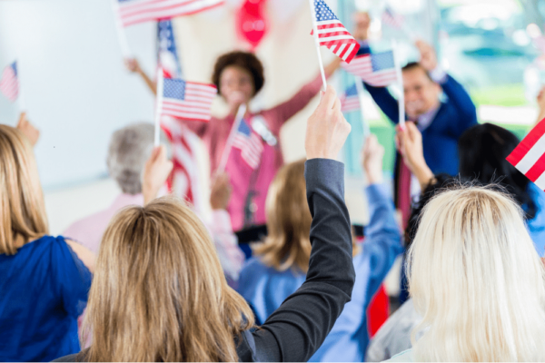 A woman stands on stage while other women wave American flags.