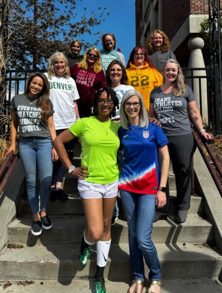 A group of women stands smiling on outdoor steps, each wearing apparel that supports women's and inclusive sports. Clothing includes slogans like "EVERYONE WATCHES WOMEN'S SPORTS." and "TRANS ATHLETES BELONG IN SPORTS," along with team shirts and athletic wear. The mood is celebratory and unified.